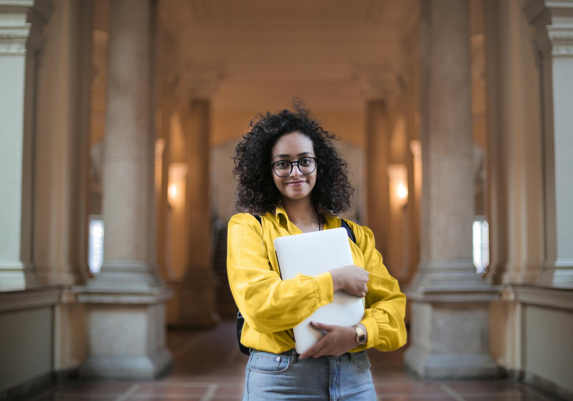 selective focus photo of smiling woman in yellow shirt and blue jeans carrying a laptop
